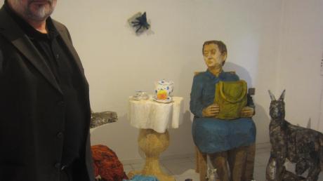 Clemens Heinl bei seiner hölzernen Sitzgruppe mit alter Frau und etlichen Tieren, wie sie in der Atelier-Galerie Oberländer zusammengestellt ist. 	