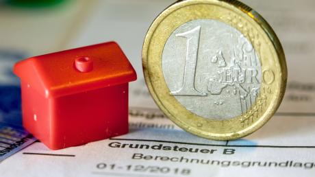 Illustration - Eine Euro-Geldmünze sowie ein Spielzeughaus stehen auf einem Abgabenbescheid für die Entrichtung der Grundsteuer.