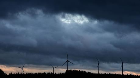Windräder stehen bei Aitrang im Allgäu unter dichten Regenwolken hinter einem Wald.