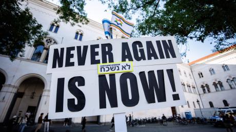 Ein Schild mit der Aufschrift „Never again is now!“ ist bei Teilnehmern einer Mahnwache gegen ein geplantes Pro-Palästina-Protestcamp vor der Ludwig-Maximilians-Universität (LMU) zu sehen.