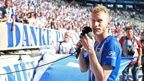 Fabian Lustenberger von Hertha BSC steht mit zwei Mikrofonen in der Hand gerührt vor der Fankurve.