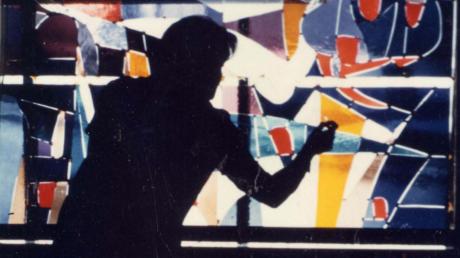 Hilda Sandtner als Silhouette bei der Arbeit an einem bunten Glasfenster in Munzingen. In der künstlerischen Gestaltung von Glas machte sie sich einen großen Namen, arbeitete aber ebenso erfolgreich mit Textil wie Stift und Papier.
