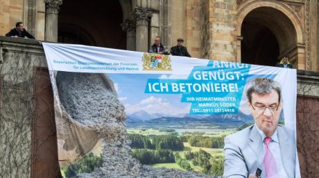Mitglieder der Grünen protestieren mit dem Plakat "Anruf genügt: Ich betoniere! - Ihr Heimatminister Markus Söder", jetzt will sich dieser auch den Flächenverbrauch eindämmen.