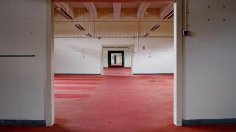 So sieht es im oberen Stockwerk der Halle 116 aus: eine endlose Betonflucht aus Fluren, Türen und wieder Fluren. Auf dem Boden ist roter, teils vergammelter und durchlöcherter Teppich verlegt. 	