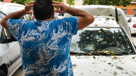 Ein Mann hält sich die Hände an den Kopf, als  er die Schäden an seinem Auto nach einem Hagelsturm sieht.